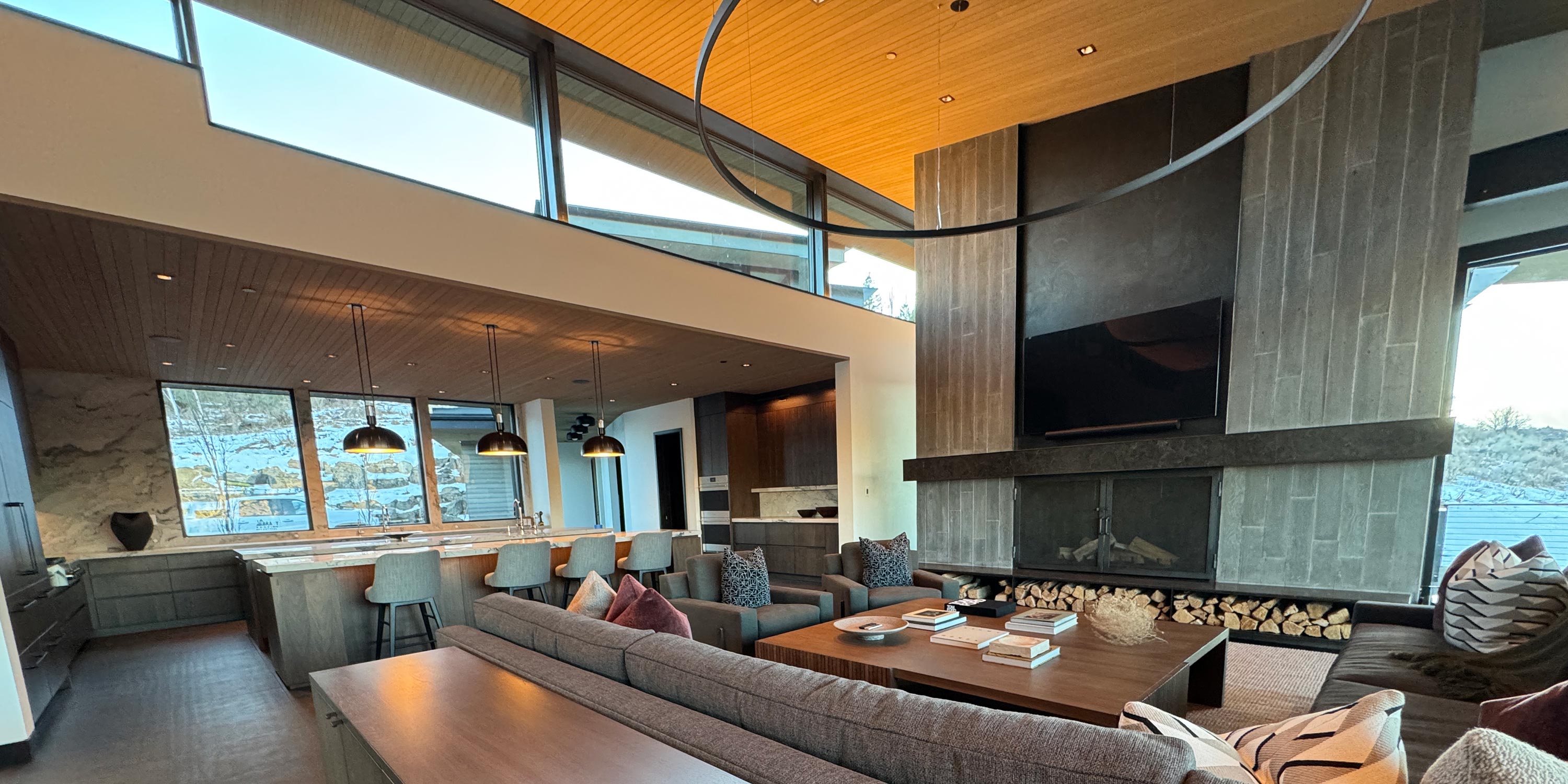 Modern open-concept living room and kitchen with large windows, wooden ceiling, and stone fireplace