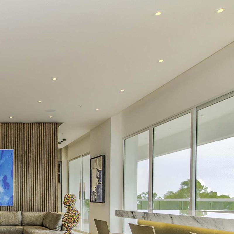 Small, round downlights installed in a white ceiling, brightening a living area with large windows and neutral d&eacute;cor.