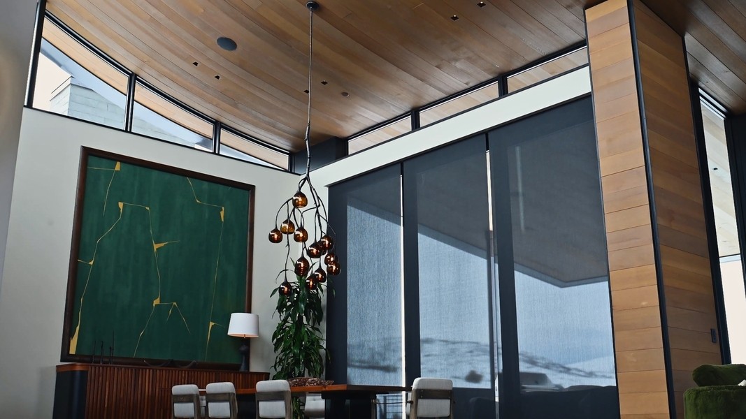 Modern dining room with tall windows and sleek motorized shades, wood ceiling, green wall art, and sculptural chandelier.
