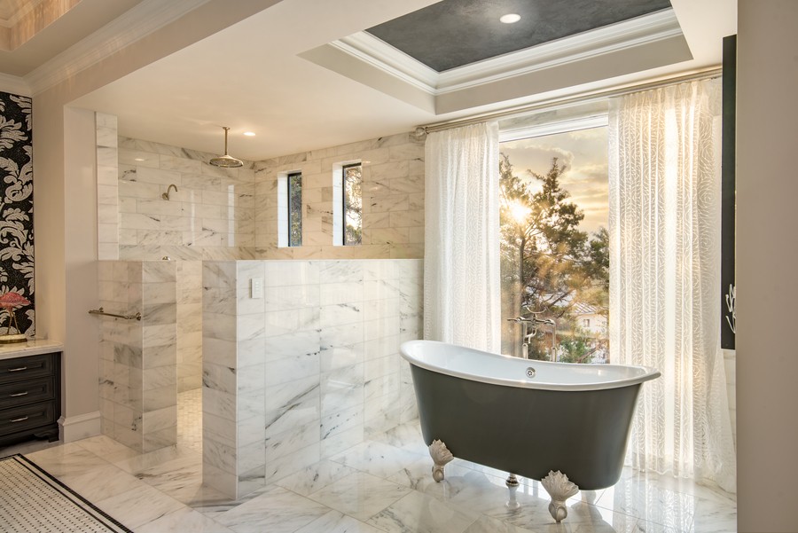 A marble bathroom with a clawfoot tub, Ketra lighting, and sunlight entering through open draperies.