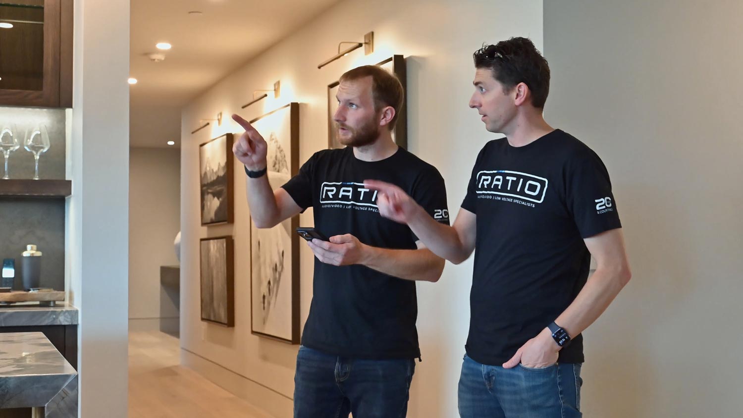 Two men are standing in a hallway, pointing and discussing while holding a smartphone. The hallway has artwork on the walls and soft lighting.
