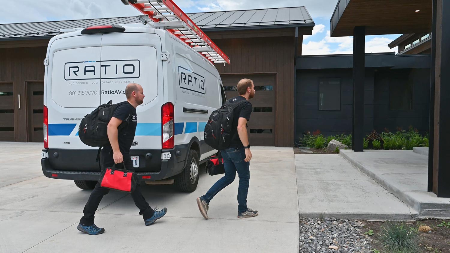 Two men with backpacks and a tool bag walk towards a house from a van parked in the driveway.