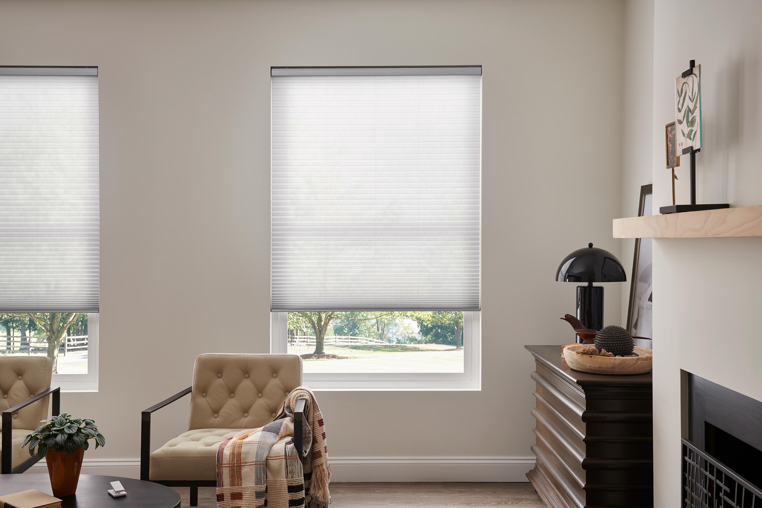 A cozy sitting area with two beige chairs, a blanket, and a potted plant in front of windows with closed blinds, creating a serene and inviting space.