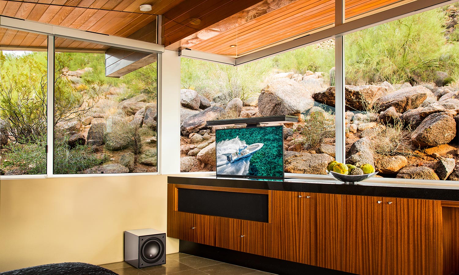 A modern living room featuring large windows with a view of rocks and plants, with a TV and speaker.