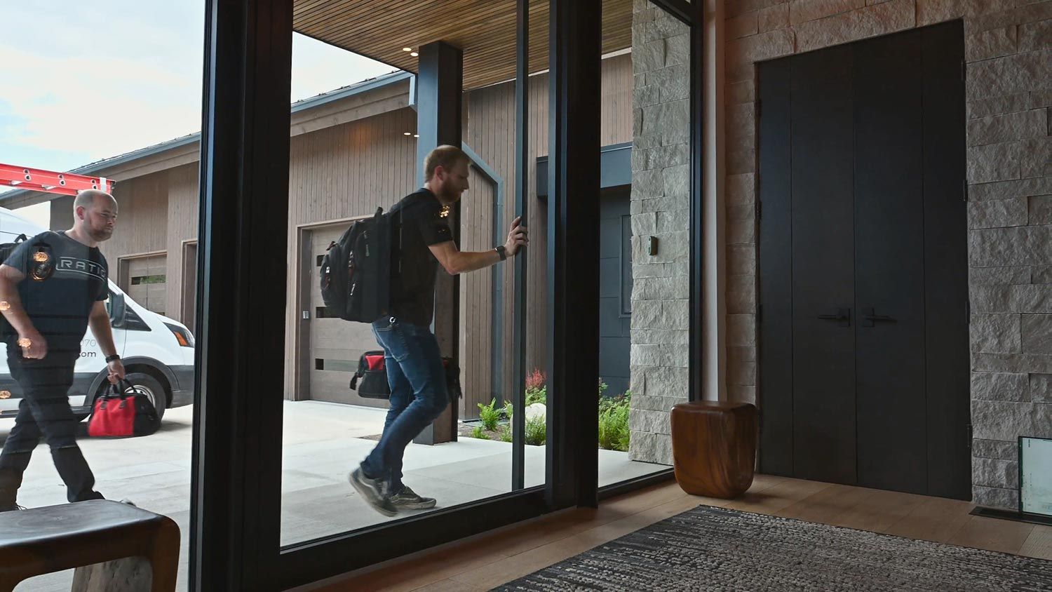 Two men carrying backpacks and a tool bag enter a modern home through a large glass door, with a white van parked outside.