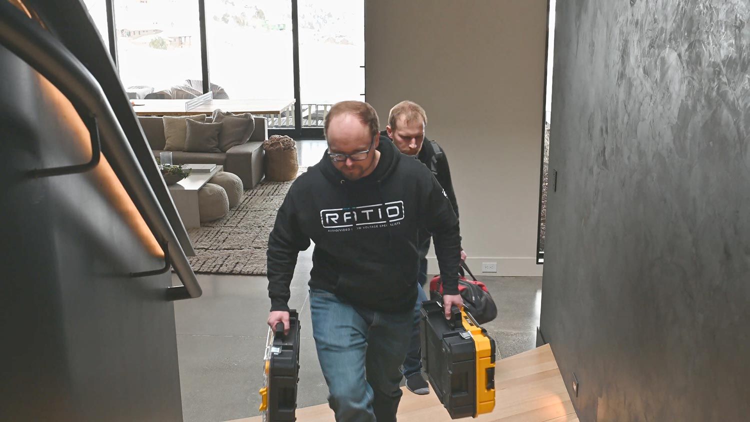 Two technicians carrying toolboxes up a staircase in a contemporary home with large windows.