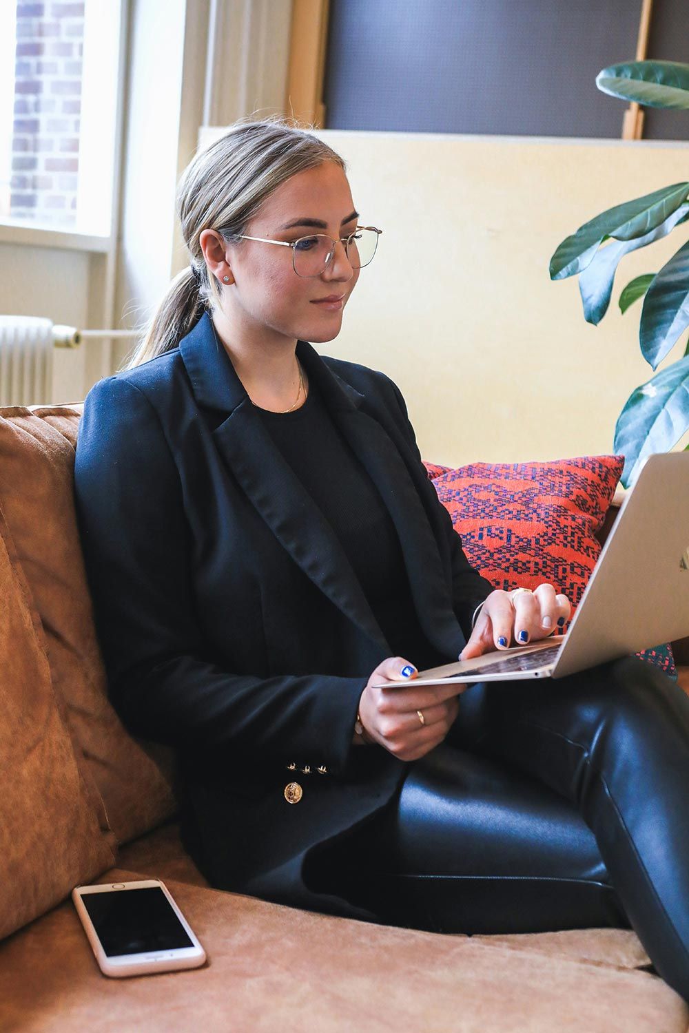 Woman in a black blazer working on a laptop with a smartphone placed on the couch next to her.